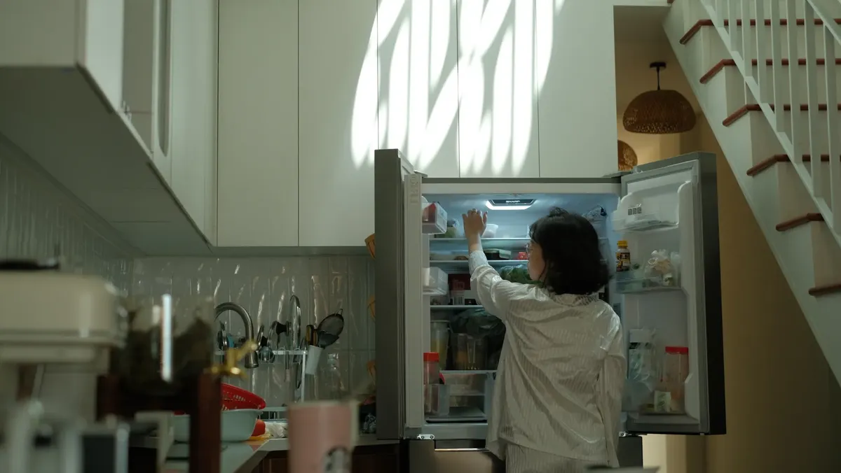 파자마를 입은 여자가 따뜻한 빛의 따뜻한 주방에서 냉장고를 확인하고 있습니다