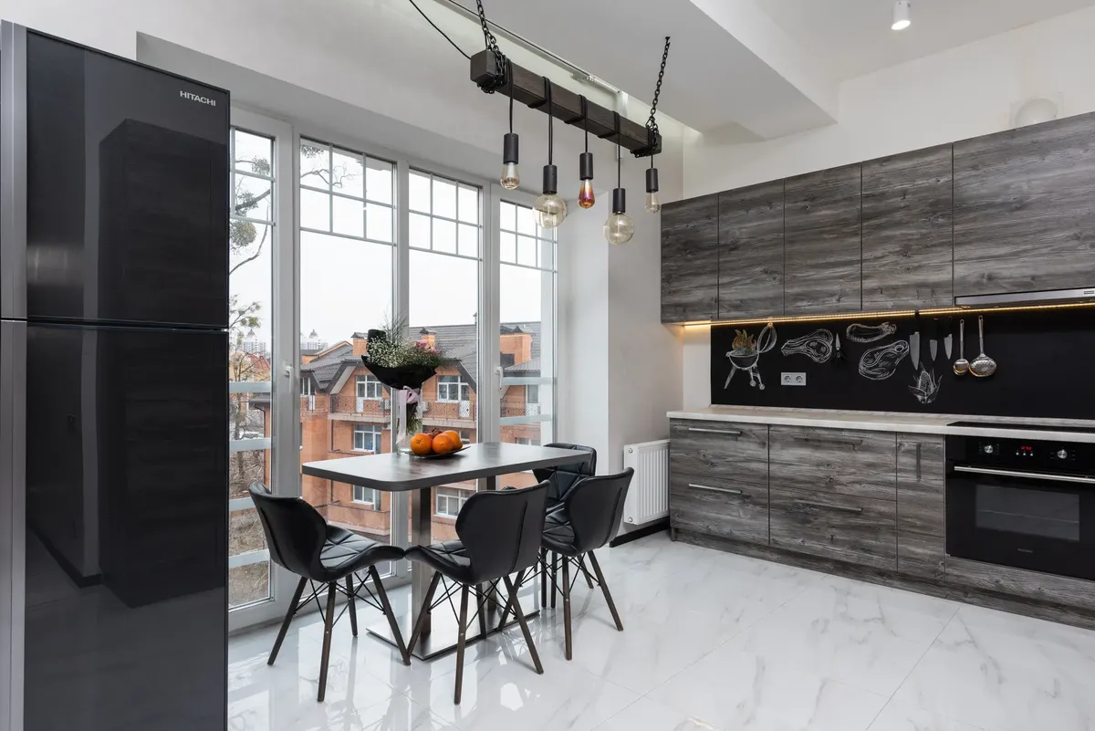 Fridge and table with chairs placed near window in stylish light kitchen with cupboards and modern kitchenware in spacious apartment