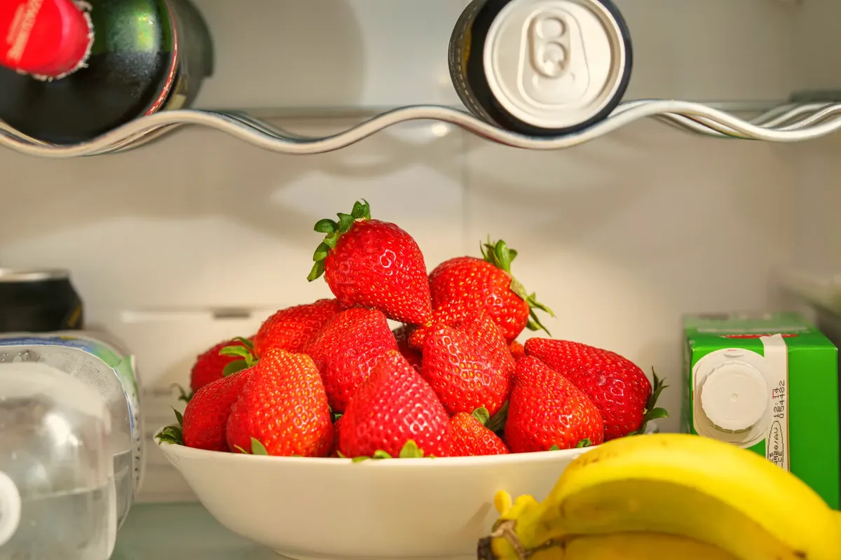 A bowl of ripe strawberries in a refrigerator, surrounded by drinks and bananas, showcasing vibrant freshness.